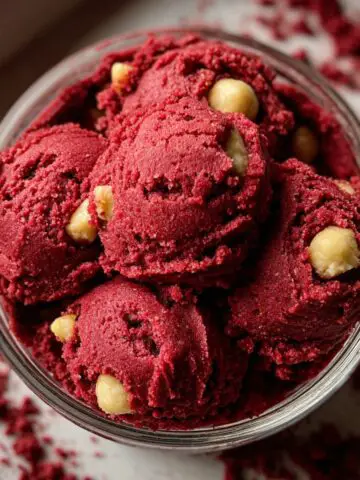 Homemade edible red velvet cookie dough scooped into a glass bowl, with a rich red color, creamy texture, and white chocolate chips throughout. Crumbs are scattered around on a light surface, giving it a fresh, homemade look.