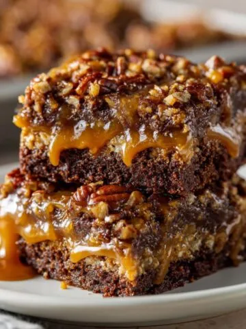 Stacked homemade Knock You Naked Bars with gooey caramel, fudgy chocolate layers, and chopped pecans on top, served on a white plate with a rustic, fresh-baked look.