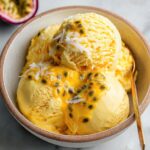 Homemade Passionfruit Ice Cream in a rustic ceramic bowl, topped with golden passionfruit pulp, coconut flakes, and a gold spoon, with half a passionfruit in the background.