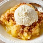 Homemade pineapple dump cake topped with a scoop of vanilla ice cream in a white bowl with a spoon.