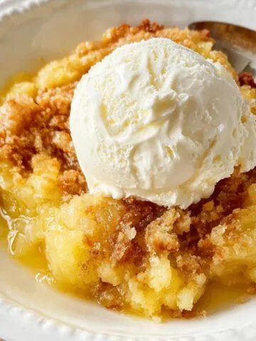 Homemade pineapple dump cake topped with a scoop of vanilla ice cream in a white bowl with a spoon.