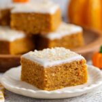 Thick, moist homemade pumpkin bars topped with white cream cheese frosting and sprinkles, served on a ceramic plate with other bars in the background.