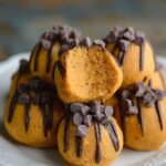 Homemade pumpkin cheesecake truffles coated in graham cracker crumbs, topped with mini chocolate chips, and one cut open to reveal smooth pumpkin cheesecake filling on a rustic plate.