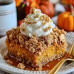 Homemade Pumpkin Crunch Cake slice on a white plate, topped with crumbly streusel, whipped cream, and caramel sauce. A gold fork rests beside it, with small pumpkins, flowers, and a coffee mug blurred in the warm autumn background.