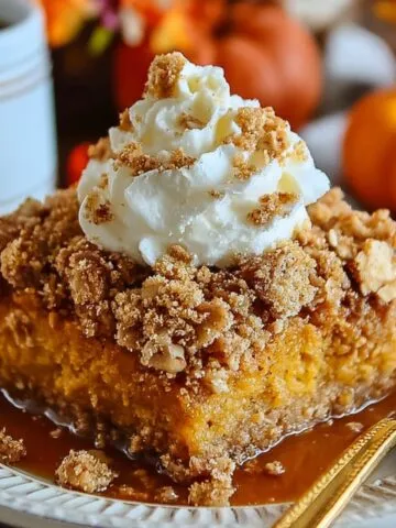 Homemade Pumpkin Crunch Cake slice on a white plate, topped with crumbly streusel, whipped cream, and caramel sauce. A gold fork rests beside it, with small pumpkins, flowers, and a coffee mug blurred in the warm autumn background.