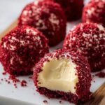 Homemade red velvet cheesecake bites on a white serving board, coated in vibrant red cake crumbs with creamy cheesecake centers, one with a bite taken out.