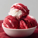 Homemade Red Velvet Ice Cream in a white bowl, showing deep red color with creamy white swirls. The bowl sits on a burgundy cloth with a soft gray background, highlighting the rich texture and vibrant contrast of the dessert.