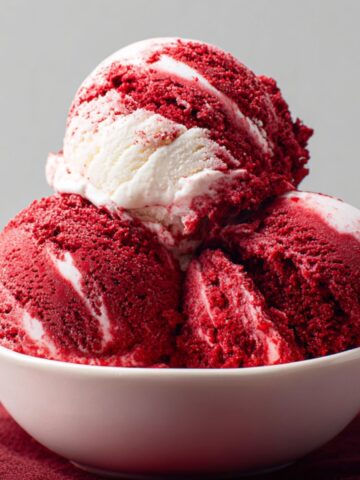 Homemade Red Velvet Ice Cream in a white bowl, showing deep red color with creamy white swirls. The bowl sits on a burgundy cloth with a soft gray background, highlighting the rich texture and vibrant contrast of the dessert.