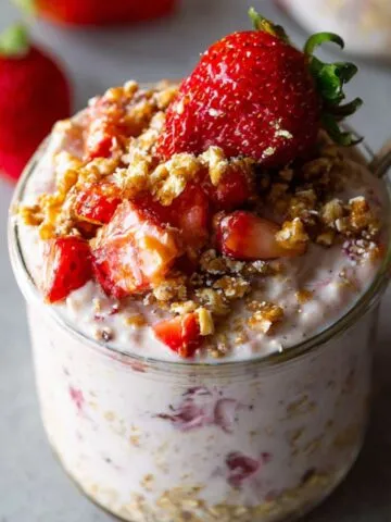 Jar of strawberry cheesecake overnight oats topped with fresh strawberries, oats, and syrup with a spoon.