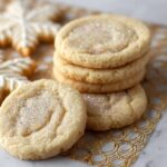 Homemade Sugar Cookie Recipe with sparkling sugar on top, stacked on a gold rack with iced snowflake cookies in the background.