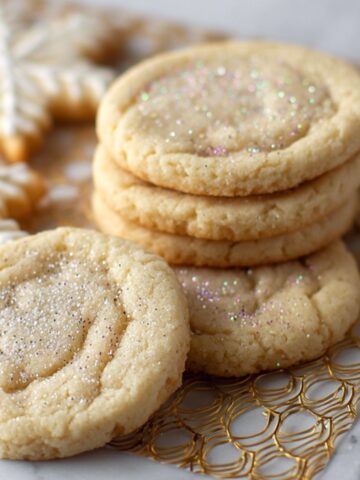 Homemade Sugar Cookie Recipe with sparkling sugar on top, stacked on a gold rack with iced snowflake cookies in the background.