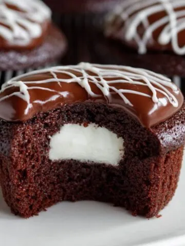 Hostess Cupcakes with cream filling, glossy ganache, and white icing swirl, with more cupcakes in the background.