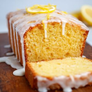 Homemade lemon drizzle cake loaf on a white plate, golden sponge sliced to show moist texture, topped with icing drizzle and lemon slices, with stacked plates blurred in the background.