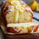 Homemade lemon cream cheese bread on a parchment-lined wooden board, topped with dripping lemon glaze and lemon slices, one slice cut to show moist crumb; whole and halved lemons blurred in the background.
