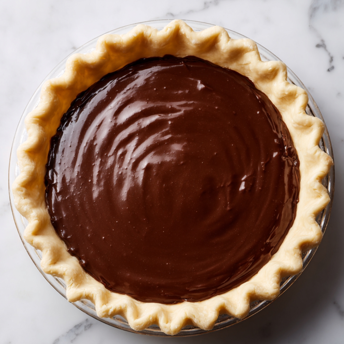 Unbaked hot fudge pie in a glass dish with crimped crust and glossy chocolate filling on a marble countertop.