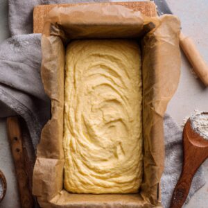 Lemon drizzle cake batter in a parchment-lined loaf tin, pale yellow with swirled ridges, surrounded by a floured wooden spoon, small flour bowl, and grey kitchen towel on a light surface.