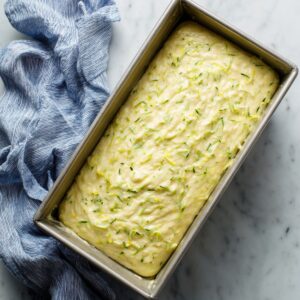 Unbaked lemon zucchini cake batter in a metal loaf pan on marble with a crumpled blue kitchen towel.
