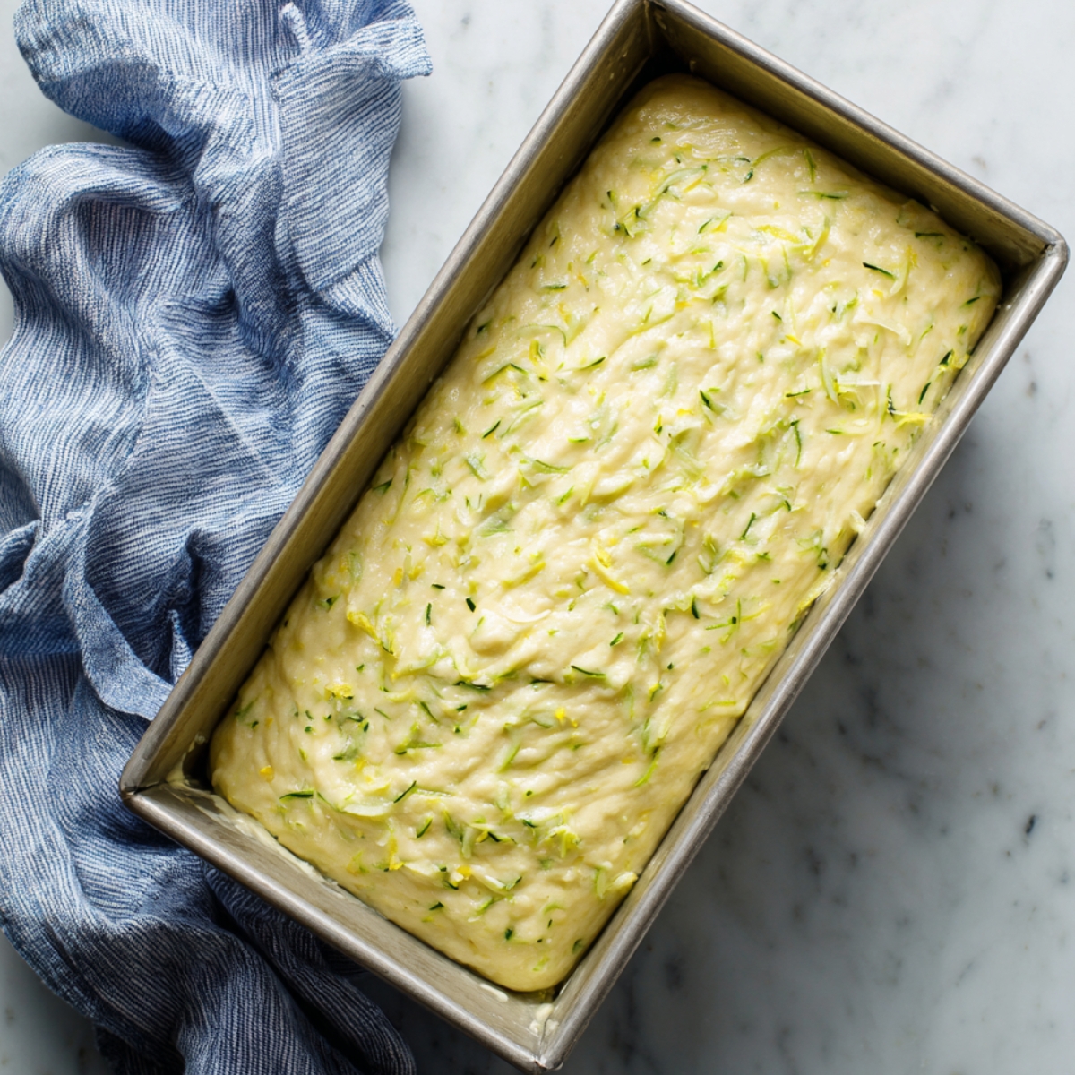 Unbaked lemon zucchini cake batter in a metal loaf pan on marble with a crumpled blue kitchen towel.