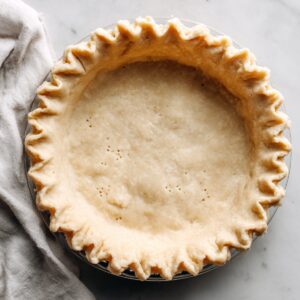 Unbaked pie crust with crimped edges in a glass dish on marble, with a beige kitchen towel nearby.