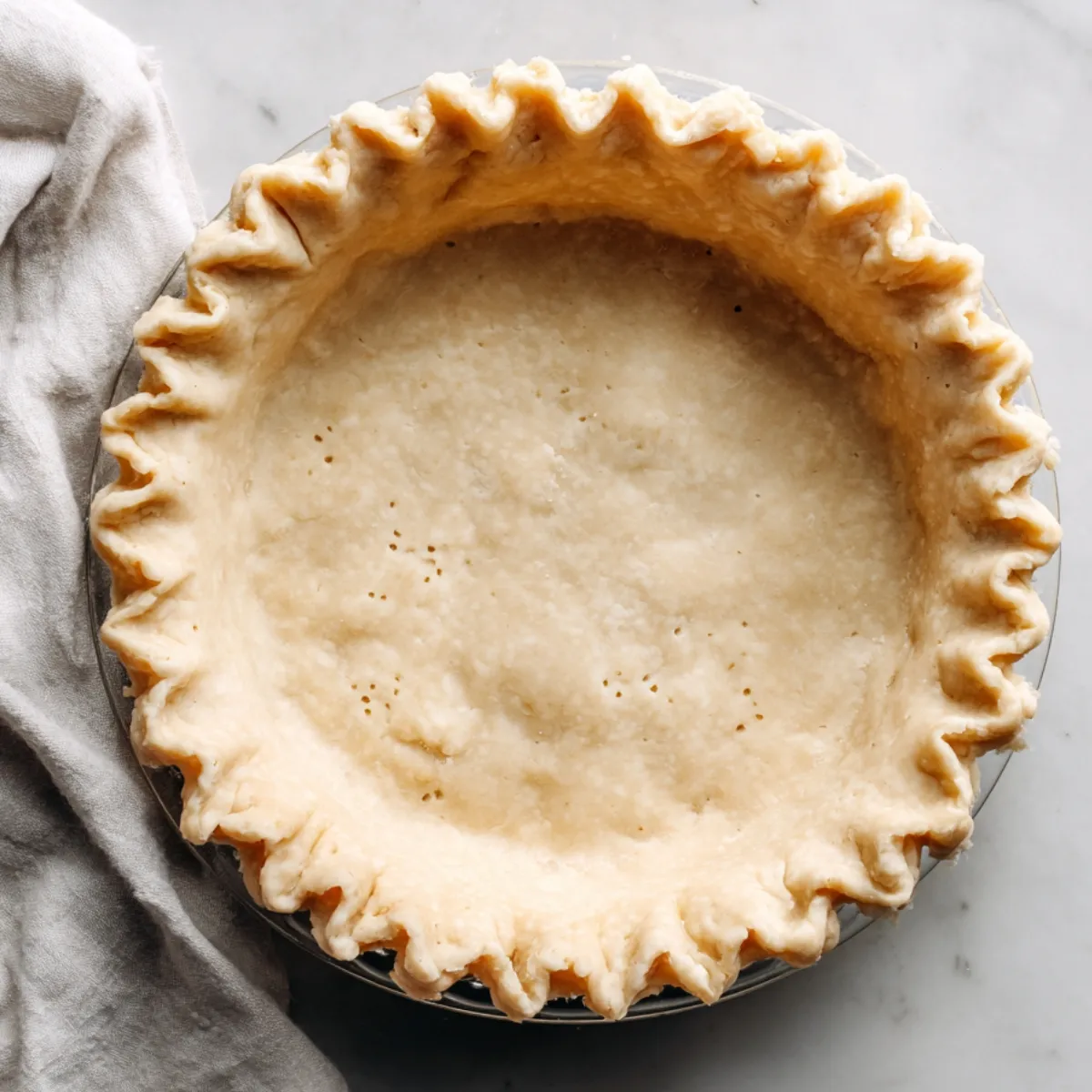 Unbaked pie crust with crimped edges in a glass dish on marble, with a beige kitchen towel nearby.
