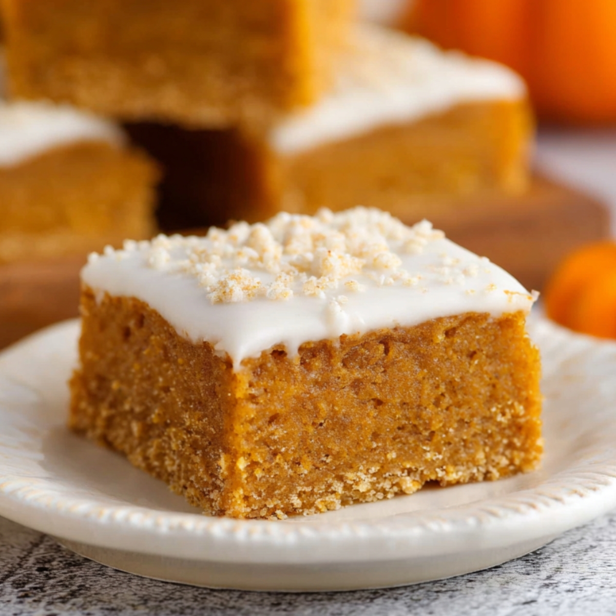 A thick homemade pumpkin bars with white icing and sugar sprinkles, served on a ceramic plate with more bars and pumpkins in the background.