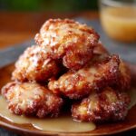 Homemade apple fritters stacked on a wooden plate, golden brown with a crisp texture, dusted with sugar and drizzled with glaze pooling on the plate.