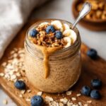 Jar of Blended Oats Recipe with peanut butter drizzle, banana slices, blueberries, and granola on a wooden board.