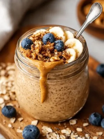 Jar of Blended Oats Recipe with peanut butter drizzle, banana slices, blueberries, and granola on a wooden board.