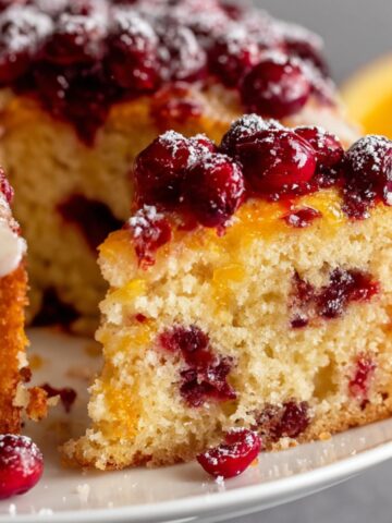 Homemade cranberry orange cake slice topped with cranberries, glaze, and powdered sugar.