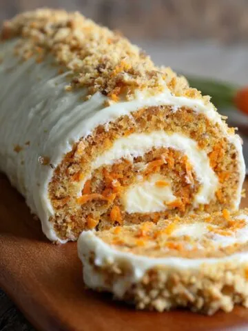 Homemade carrot cake roll filled with cream cheese frosting, sliced to reveal its spiral layers of moist spiced carrot cake and creamy filling, topped with frosting and sprinkled with golden crumbs on a wooden board.