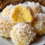 Homemade pineapple coconut balls on a white plate, one with a bite showing the soft yellow center.