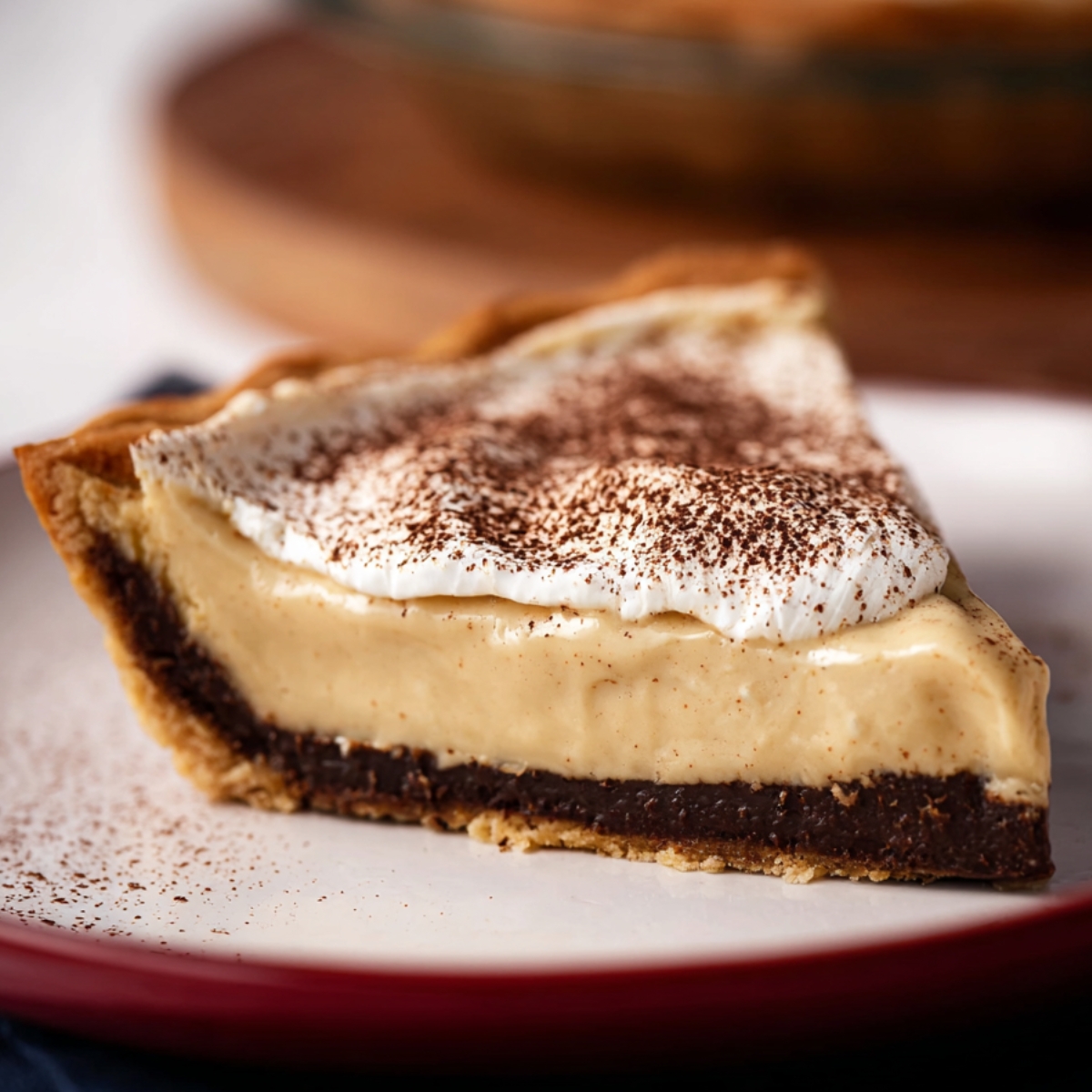 Slice of coffee custard pie with crumb crust, rich chocolate layer, creamy coffee filling, whipped cream, and cocoa dusting on a white plate.