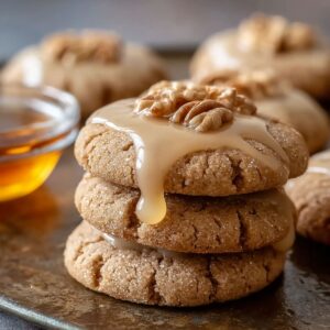Stack of homemade maple cookies topped with maple glaze and pecans, with glaze dripping down the sides, set on a rustic tray beside a small bowl of maple syrup.