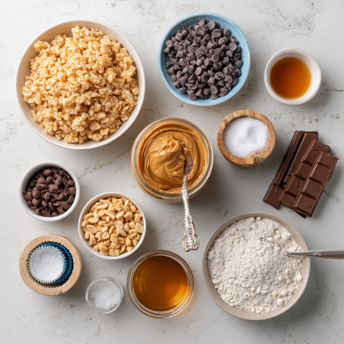 Overhead view of ingredients for homemade Chocolate Peanut Butter Rice Krispie Cups, including puffed rice cereal, peanut butter, chocolate chips, chopped peanuts, flour, sugar, honey, vanilla, sea salt, and cupcake liners, neatly arranged in bowls on a light marble surface.