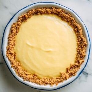 Atlantic Beach Pie in a white pie dish with a blue rim, showing a smooth pale yellow citrus filling and a crumbly golden-brown saltine crust on a marble surface.