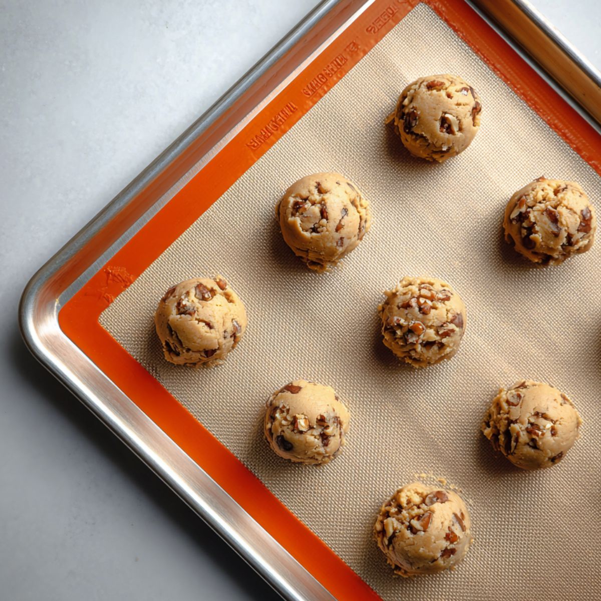 Homemade maple cookie dough balls with chopped nuts on a silicone baking mat-lined sheet pan, ready for baking.