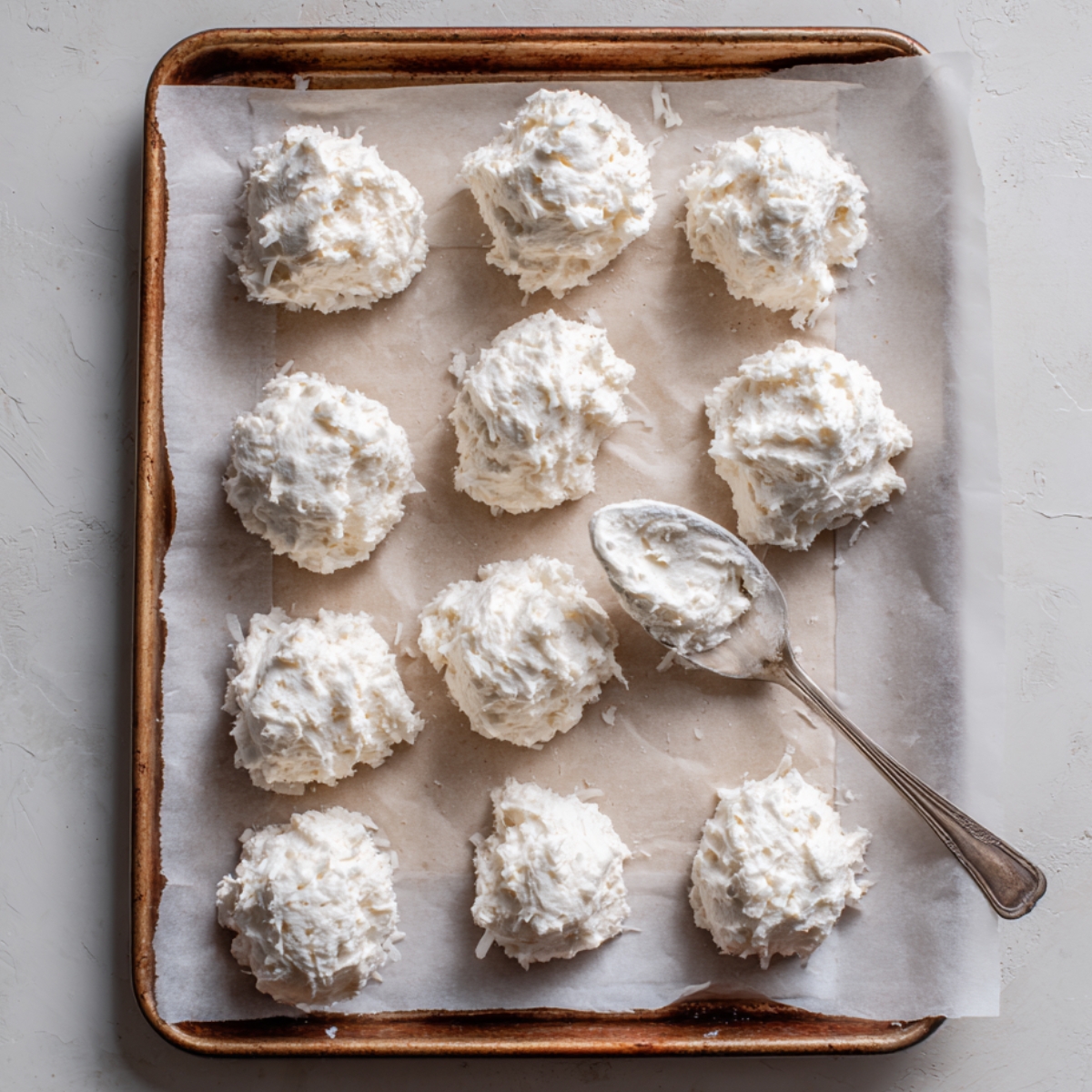 Raw coconut meringue cookie mounds on a parchment-lined baking sheet with a spoon resting beside them, rustic and homemade in appearance.