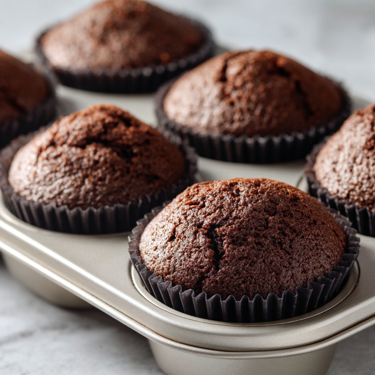 Freshly baked chocolate cupcakes in a muffin tin, warm and fluffy with cracked tops and rich brown color, still in dark paper liners on a marble countertop.