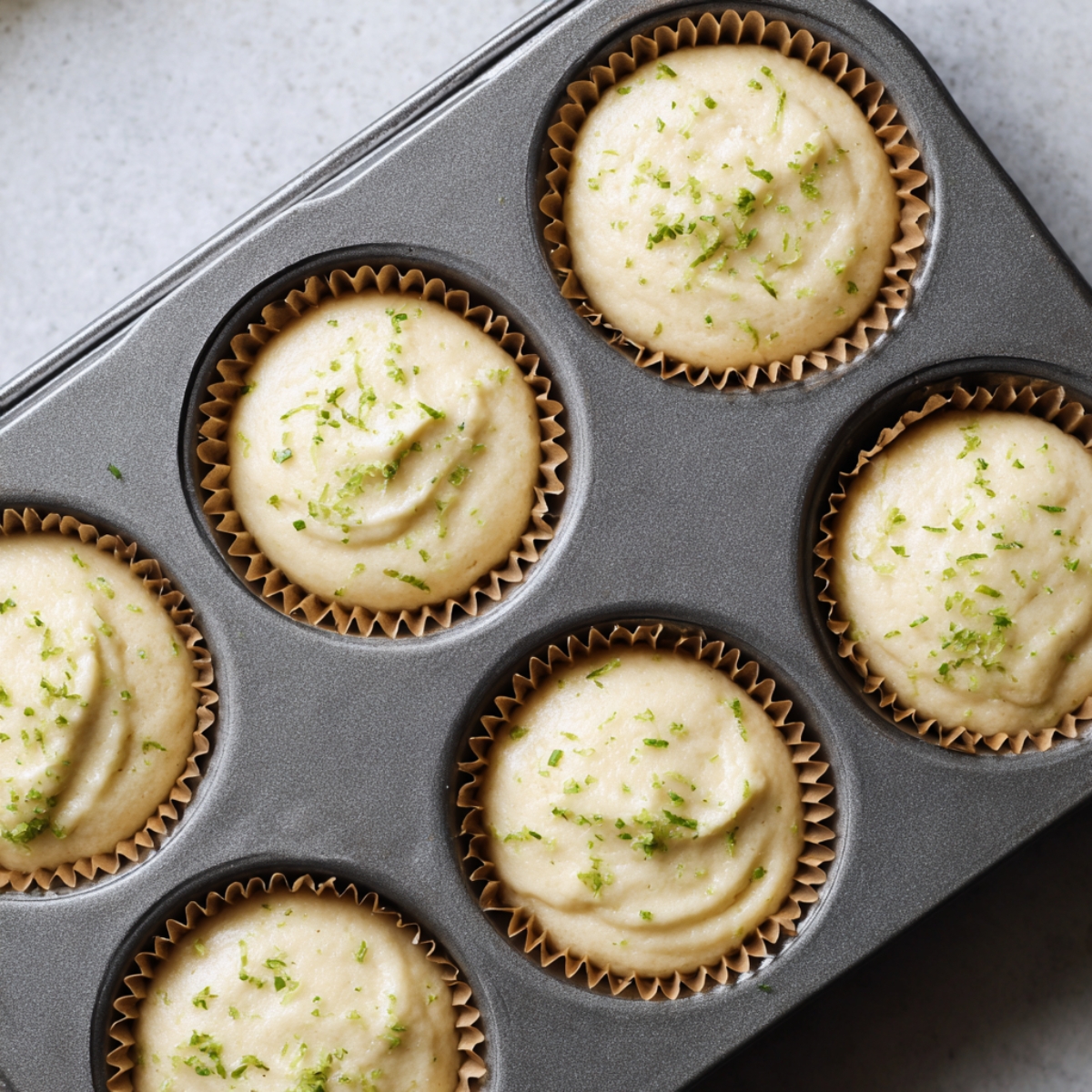 Overhead view of key lime cupcake batter in brown liners inside a muffin tin, topped with lime zest and ready to bake, lit by soft natural light.