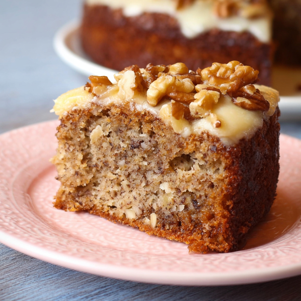 Close-up of a moist slice of homemade banana walnut cake on a pink plate, topped with creamy frosting and walnuts, with the rest of the cake blurred in the background.