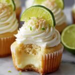 Best Key Lime Pie Cupcakes with creamy lime filling, fluffy white frosting, and a lime slice on top. Sprinkled with graham crumbs and zest, the cupcake has a bite showing its soft golden cake on a marble surface with limes blurred in the background.