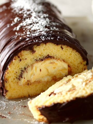 Homemade Boston cream roll cake with soft yellow sponge, creamy vanilla filling, and glossy chocolate ganache, dusted with powdered sugar on top.