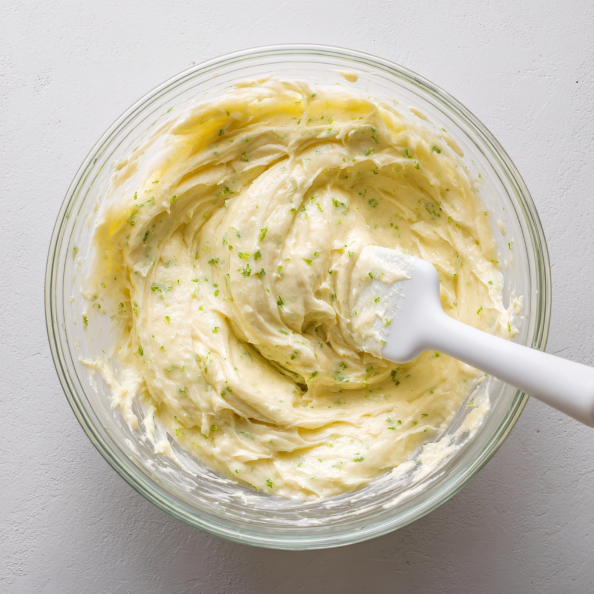 Glass bowl filled with creamy key lime cupcake batter, speckled with lime zest, and a white spatula resting inside on a light kitchen surface in natural light.