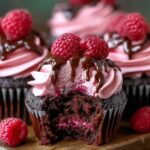 Chocolate Raspberry Cupcakes topped with pink raspberry buttercream, fresh raspberries, and a drizzle of melted chocolate, with a bite taken out to show the moist chocolate cake and raspberry filling inside.