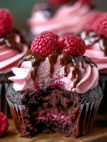 Chocolate Raspberry Cupcakes topped with pink raspberry buttercream, fresh raspberries, and a drizzle of melted chocolate, with a bite taken out to show the moist chocolate cake and raspberry filling inside.