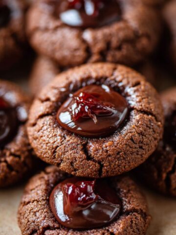 Homemade chocolate thumbprint cookies topped with glossy ganache and a bit of red jam, showing cracked edges and rich brown texture under warm natural light.