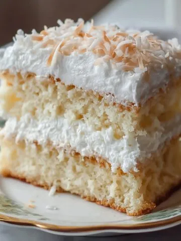 A slice of homemade coconut cloud cake with fluffy yellow layers, creamy white frosting, and shredded plus toasted coconut on top, served on a plate.