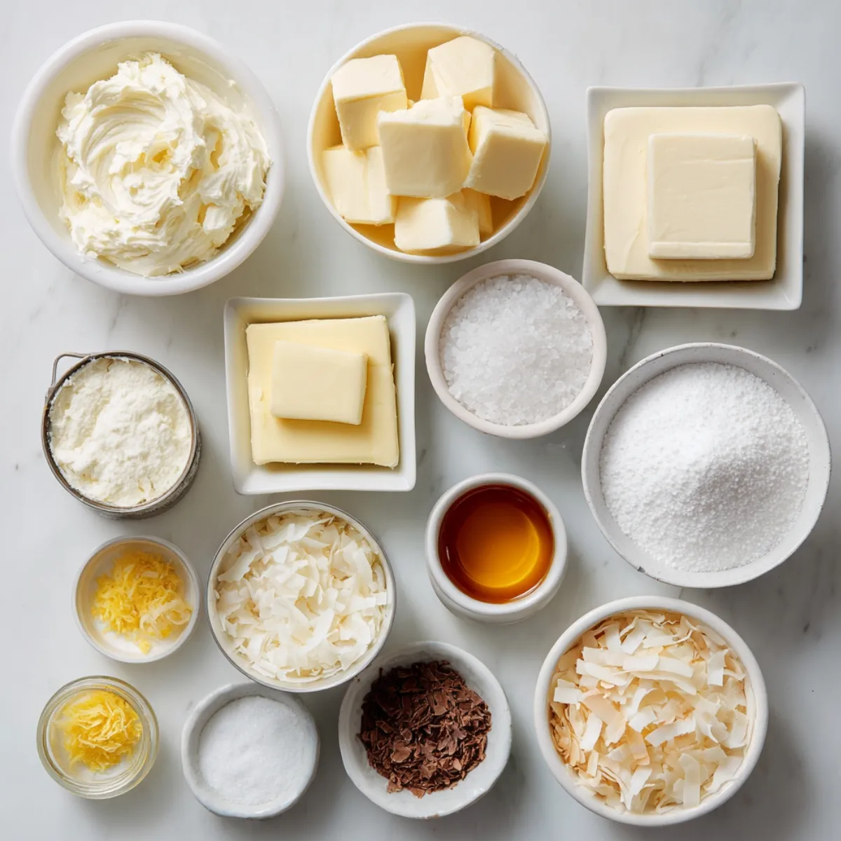 A variety of ingredients for Coconut Cloud Cake on a marble countertop: whipped butter, cubes of cold butter, a block of butter, granulated sugar, sea salt, vanilla extract, lemon zest, shredded coconut, toasted coconut flakes, chocolate shavings, and powdered sugar, all neatly arranged in bowls.