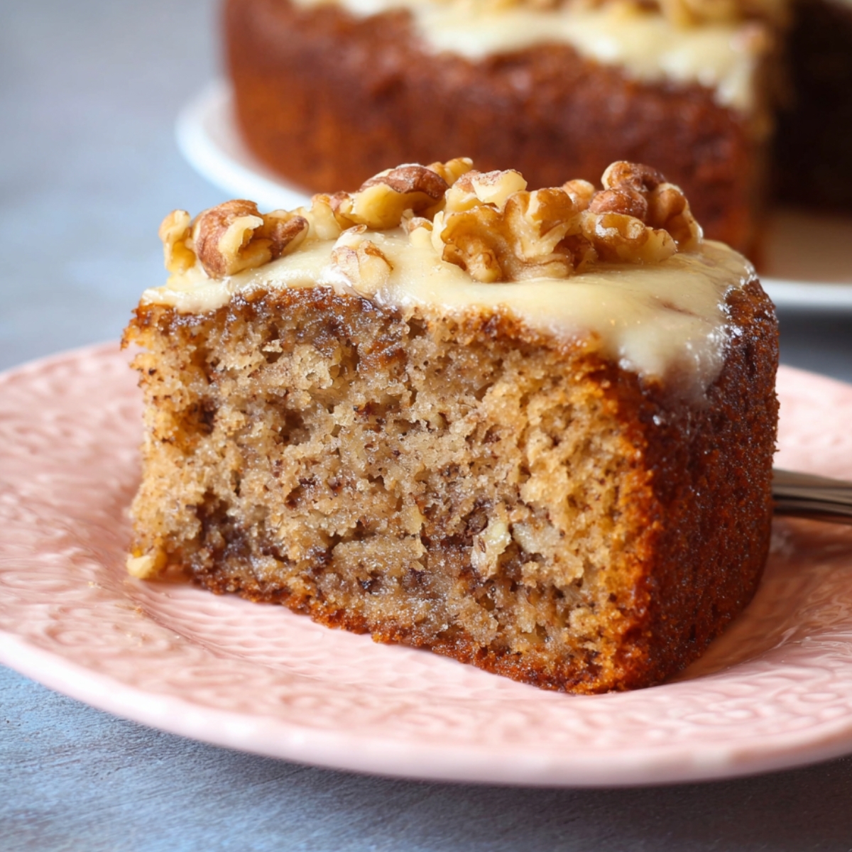 Easy Moist Banana Walnut Cake Recipe slice on a pink plate, topped with creamy frosting and chopped walnuts, with the rest of the cake blurred in the background.