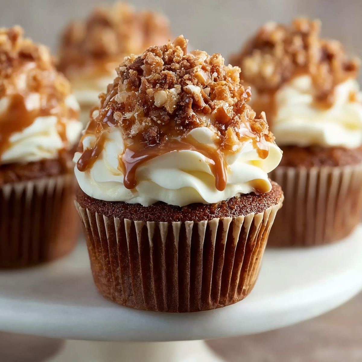 Easy Toffee Crunch Cupcakes Recipe topped with caramel buttercream, drizzled caramel, and crushed toffee bits on a white cake stand, in warm natural light.